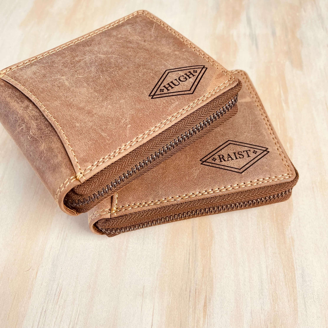 Two brown leather wallets with embossed names on a wooden surface.